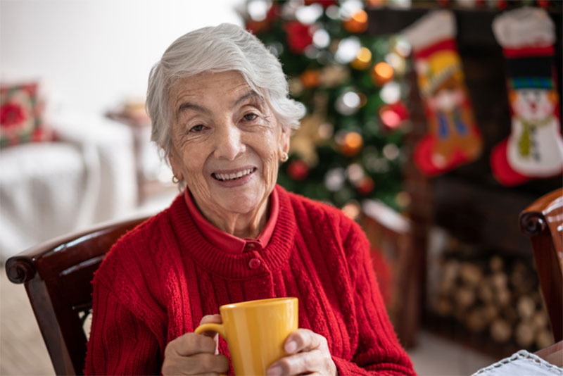 A senior woman enjoys a cup of coffee while sitting by her Christmas tree. When visiting older loved ones this holiday season, know the warning signs to watch for that help may be needed.